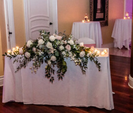 White table with white and pale pink rose wedding flowers, greenery, and candles, in a warmly lit indoor setting.