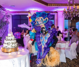 Person in a vibrant purple and blue feathered costume dances at an indoor wedding with cake and formally dressed guests nearby.
