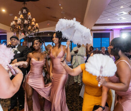 Bridesmaids in pink dresses dance with feathered fans and a parasol at a lively wedding reception in a decorated banquet hall.