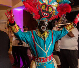 Person in colorful costume and feathered headdress poses with arms raised; brass musicians play in the background.