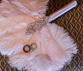 Wedding rings on a pink feather fan with jeweled handle, set atop a patterned brown and gold surface.