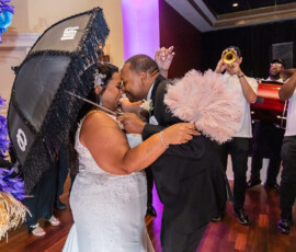 Bride and groom dance at their wedding, holding an umbrella and feather fan, with a lively brass band in a festive indoor setting.