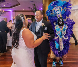 Bride and groom dance at their wedding as a guest in a blue feathered costume approaches; guests watch in the festive setting.