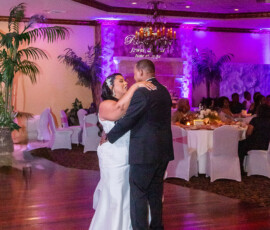Bride and groom share their first dance in a softly lit ballroom with purple lighting and guests at decorated tables.