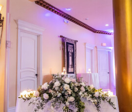 Elegant wedding table with white flowers, greenery, and candles; ornate doors, gold column, framed picture in warm lighting.