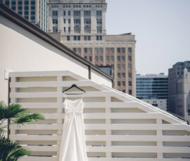 White wedding dress on a black hanger against a white fence, rooftop setting with city buildings in the background.