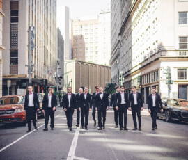 Nine men in suits and bow ties walk down a city street lined with tall buildings and parked cars in bright daylight.