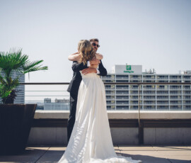Bride and groom embrace on a rooftop with city buildings and clear sky; she wears white, he a black suit.