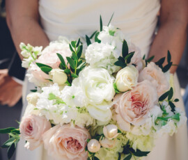 Bride in a white dress holding a bouquet of pale pink and white roses, ranunculus, and greenery.