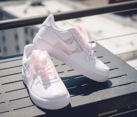 White sneakers with pink glitter Nike swooshes and laces on a dark outdoor surface, cityscape blurred in the background.
