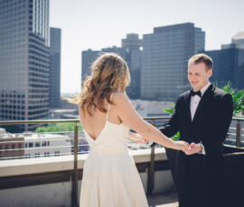 Bride and groom in wedding attire hold hands on a rooftop with city skyscrapers behind them, smiling in sunlight.