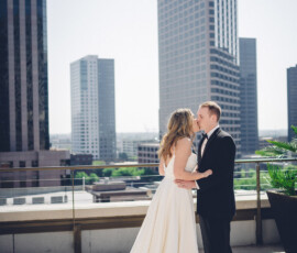 Bride and groom embrace on a sunny rooftop, gazing at each other with city buildings in the background.