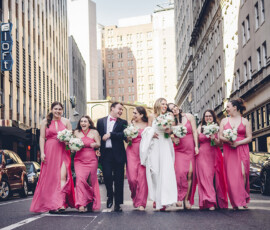 Bride, groom, and six bridesmaids in pink dresses walk happily down a city street with bouquets, tall buildings, and cars.