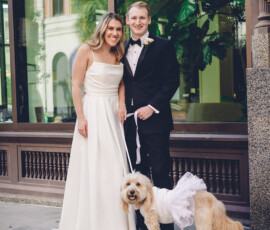 Bride and groom smile on a sidewalk with their white dog in a tutu; large window and green plants behind them.
