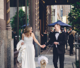 Bride and groom smile at each other, walking hand in hand outside a hotel with their dog in a frilly white outfit.