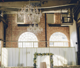 Wedding ceremony setup with rows of chairs, floral arch, and table in an industrial space with brick walls and arched windows.