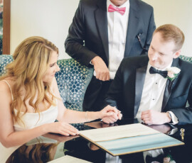 Bride and groom smile as they sign their wedding document, with a suited person in a pink bow tie standing behind them.