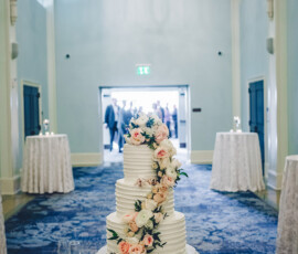 Four-tier white wedding cake with pink and white flowers on a round table in an elegant hall with blue carpet and high ceilings.