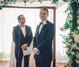 Groom in black tuxedo smiles under floral arch as officiant with red bow tie and microphone stands beside him holding papers.