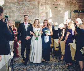 Bride with bouquet walks down aisle between two adults, guests seated in sunlit room with brick walls and chandelier.
