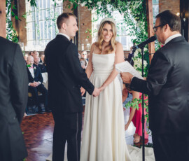 Bride and groom smile and hold hands at an indoor wedding ceremony, with officiant, guests, greenery, and flowers present.