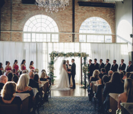 Bride and groom hold hands under a floral arch at their sunlit wedding, with guests and bridal party looking on.