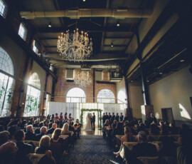 Elegant wedding ceremony in a grand hall with chandeliers, large windows, and guests seated on both sides facing the couple.