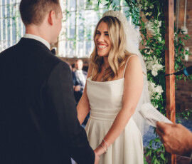 Smiling bride in white dress and veil holds hands with groom at indoor wedding, guests and greenery-decorated windows behind.