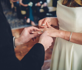 Couple in formal attire exchanging wedding rings, close-up focus on their hands.