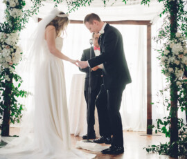Bride and groom exchange vows under a floral arch, holding hands as sunlight streams through tall windows behind them.