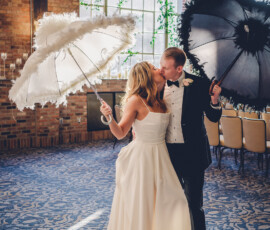 Bride and groom kiss indoors beneath sunny arched windows, holding white and black feathered parasols.