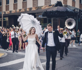 Newlyweds walk hand-in-hand down a city street with a white parasol, followed by a brass band and joyful crowd.
