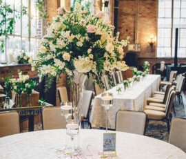 Round table with floral centerpiece, candles, and cards in a sunlit venue with banquet tables, large windows, and brick walls.