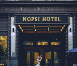Bride and groom embrace outside the NOPSI Hotel, elegant building and lights shining in the background.