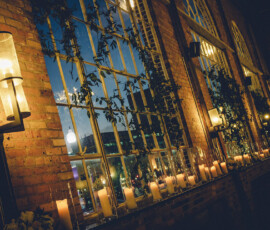 Lit candles on a windowsill and wall sconces cast a warm glow in a room with arched windows, brick walls, and leafy vines.