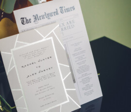 Wedding invitation and personalized newspaper program on a reflective black surface with a decorative vase behind.
