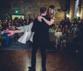 Man in suit lifts smiling woman in white dress and pink shoes as guests at tables watch and cheer at formal indoor event.