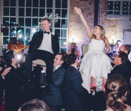 Bride and groom lifted on chairs by guests at a lively wedding reception with smiling faces and exposed brick walls.