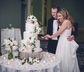 Bride and groom smiling while cutting a white tiered wedding cake with pink and white flowers at their decorated reception hall.