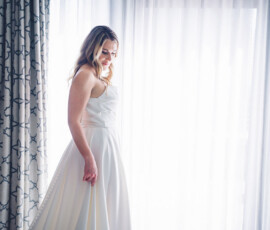 Woman in white sleeveless dress by window with sheer curtains, softly illuminated, looking down. Patterned drapes frame scene.