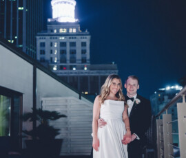 Smiling bride in white gown and groom in black tuxedo on rooftop at night with glowing city building behind them.