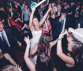 Woman in white dress dancing joyfully amid lively party crowd with glowing sticks on the dance floor.