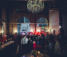 Guests dance at a dimly lit indoor wedding with a band, arched windows, chandelier, and candlelit tables with flowers.