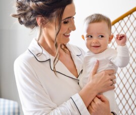 A woman in white pajamas holds a smiling baby indoors by a wooden railing, sharing a warm, tender moment.