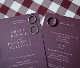 Burgundy wedding invitations with rings on a checkered tablecloth, featuring Abby P. Bender and Patrick C. Sullivan’s event details.