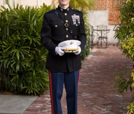 Man in military uniform on brick walkway holding white hat, smiling with greenery and building—Louisiana wedding inspiration.