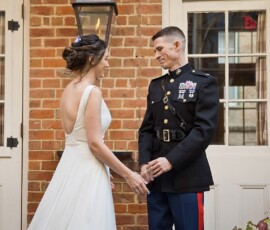 Bride in white dress holds hands with man in military uniform, smiling in front of brick wall and white doors at New Orleans wedding.