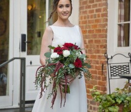 Bride in white dress smiling outside with red and white bouquet near brick wall and glass doors at New Orleans wedding.