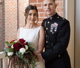 Bride in white dress with bouquet and groom in military uniform smile by brick wall at elegant New Orleans wedding.