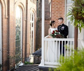 Bride and groom on white balcony in New Orleans; groom in military uniform, bride with bouquet, brick buildings behind.